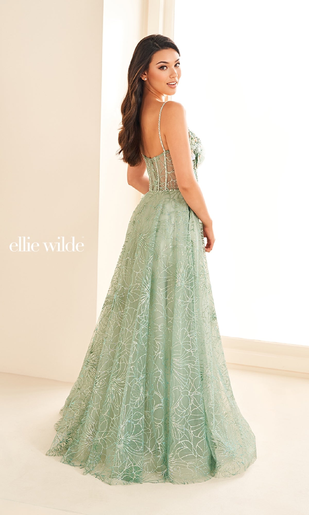 A woman stands in a brightly lit room wearing the Ellie Wilde EW36218, a light green, floor-length prom dress featuring lace details, spaghetti straps, and a sheer corset bodice.